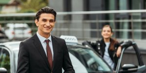 A smiling businessman in a suit and tie stands next to a taxi