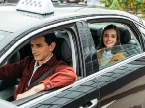 A man driving a car with a smiling woman passenger, both looking happy and engaged with the camera.